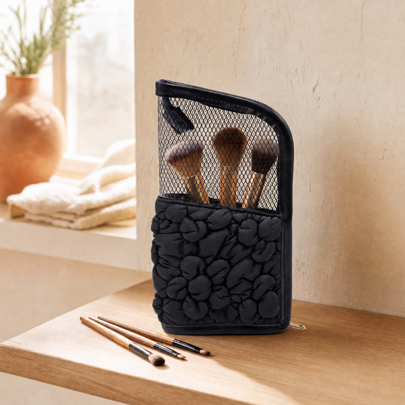 A black makeup brush pouch with a textured floral quilted bottom and a mesh upper pocket holding several makeup brushes, set on a wooden table in a warm, sunlit room.