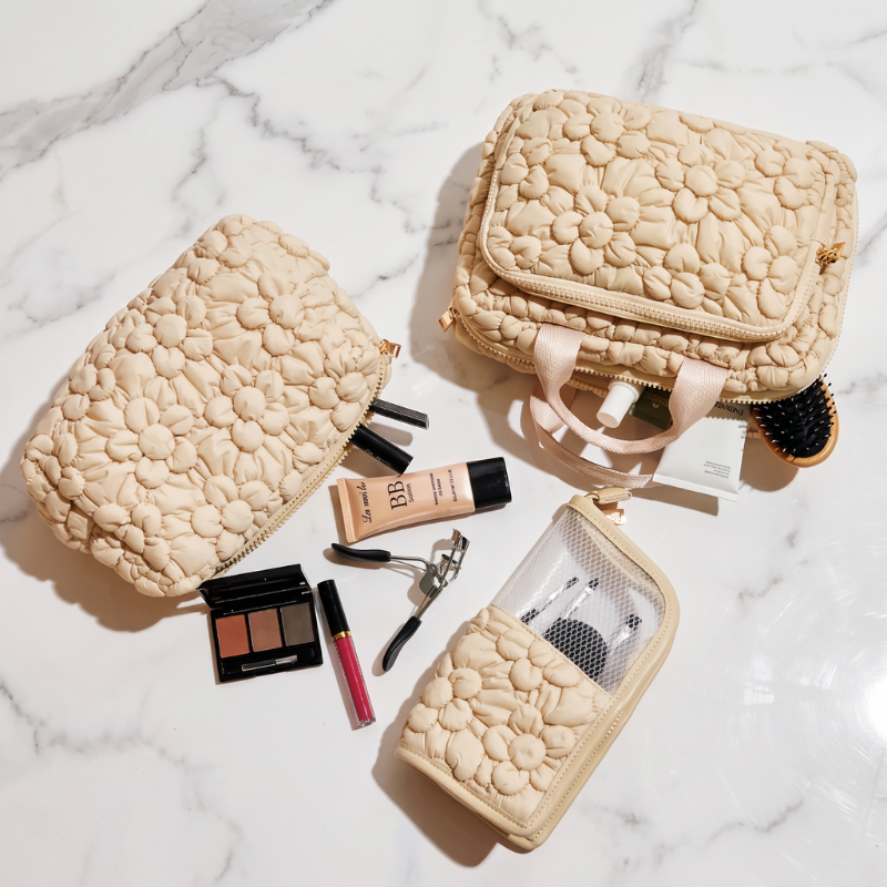 Three beige quilted floral-patterned cosmetic bags arranged on a white marble surface with makeup items scattered around them, including an eyeshadow palette, lip gloss, mascara, and a hairbrush.